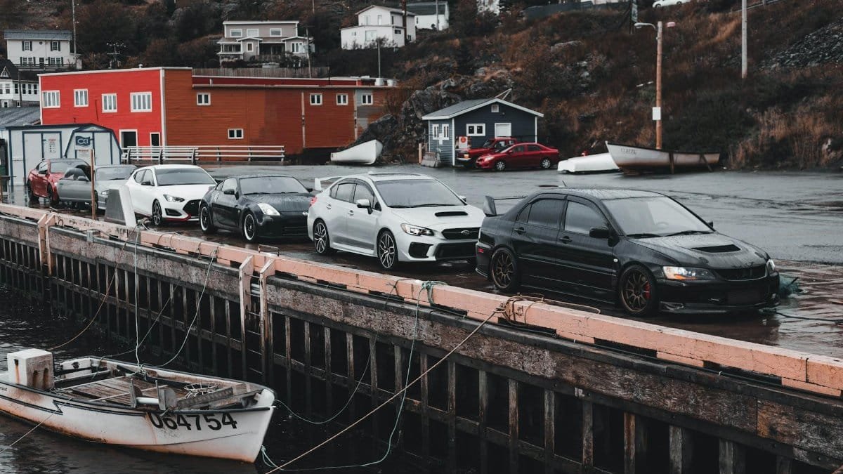 A lineup of parked cars by the waterfront in a quaint seaside town, showcasing colorful buildings and boats.