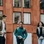 Group of cheerful multiethnic friends in casual outfit smiling near modern building of university with flag