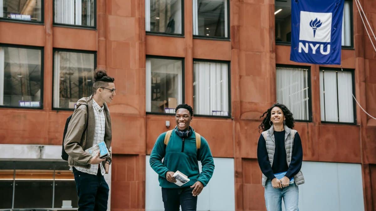 Group of cheerful multiethnic friends in casual outfit smiling near modern building of university with flag