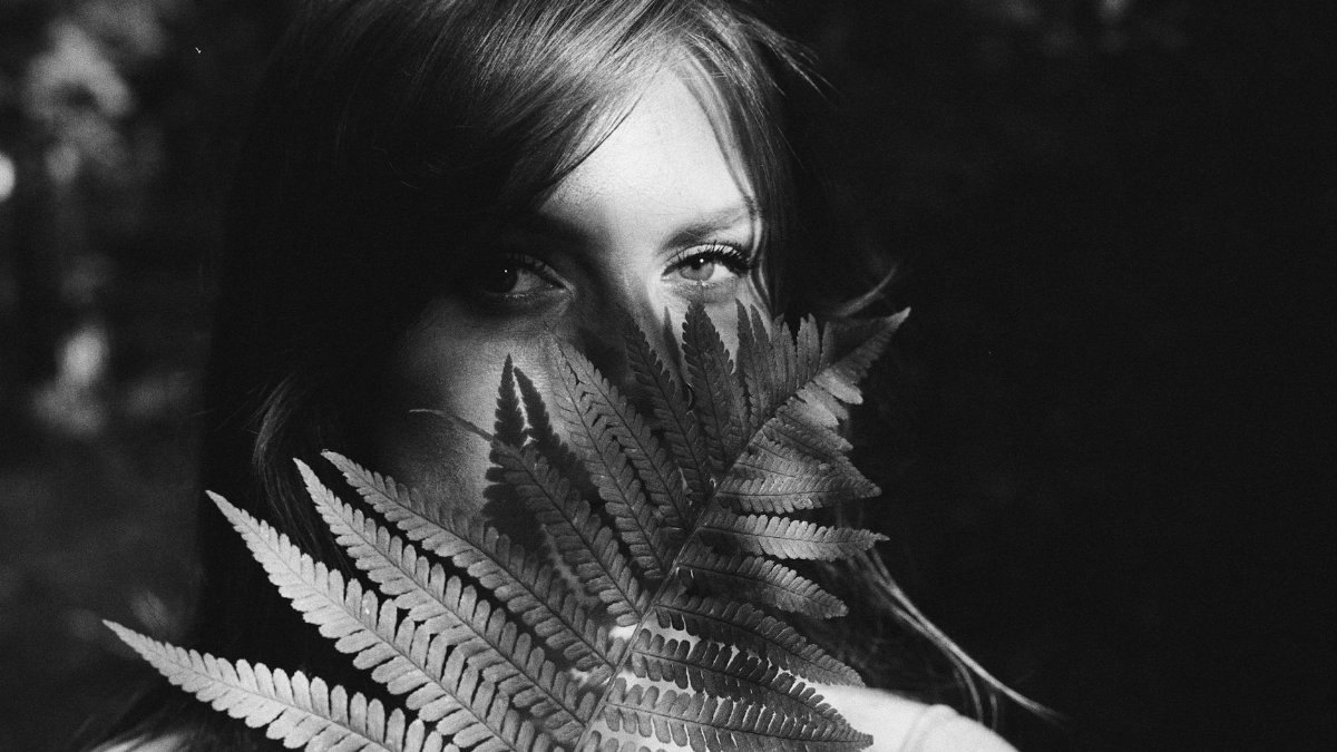 Black and white portrait of a woman partially hidden by fern leaves, creating a mysterious, artistic mood.
