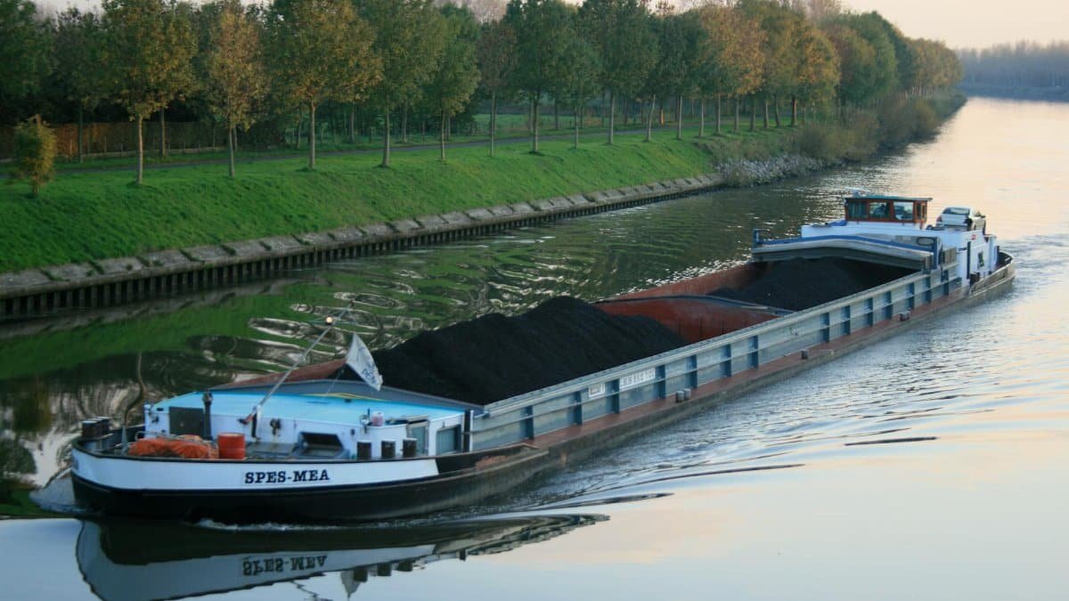 Large cargo barge carrying coal along a tranquil canal lined with lush trees during sunset.