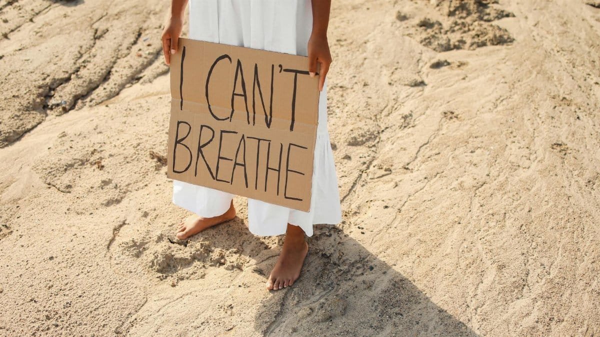 A woman stands barefoot on sand holding a protest sign stating 'I can't breathe'.