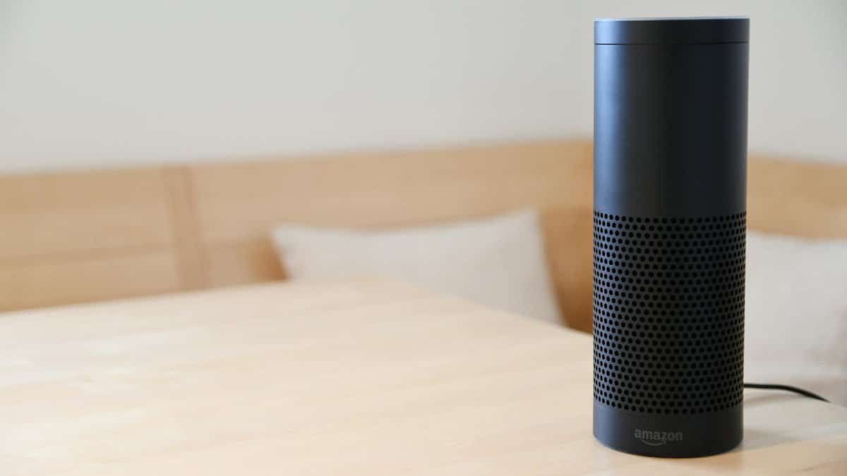A black smart speaker resting on a light-colored wooden table in a cozy indoor setting.