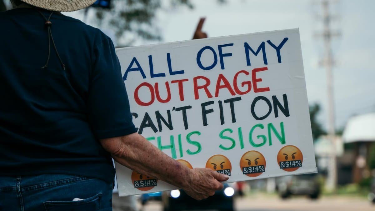 Person at a protest holding a sign expressing their outrage. Outdoors setting.