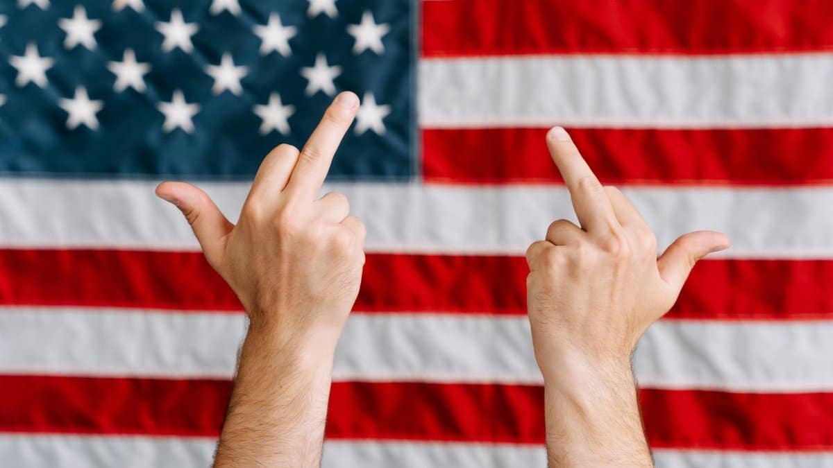 Hands displaying a rebellious gesture in front of the American flag, symbolizing defiance.