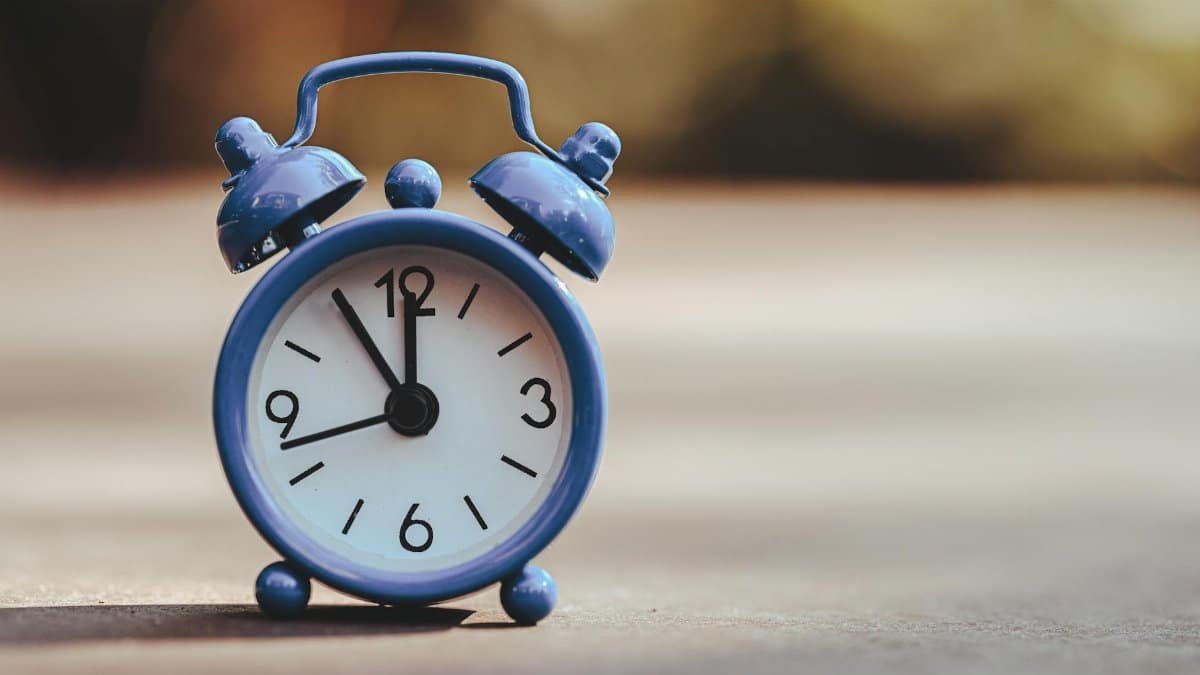 A classic blue alarm clock placed outdoors, displaying five minutes to twelve.