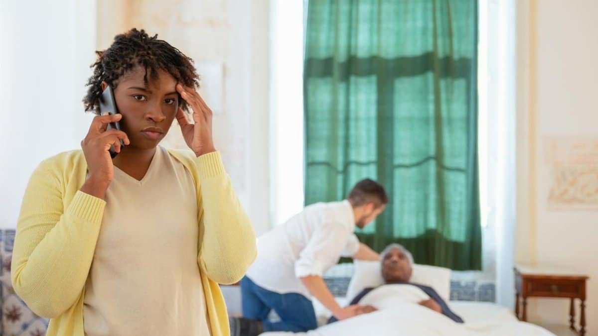 A woman on the phone looks worried as a caregiver assists an elderly person in bed.