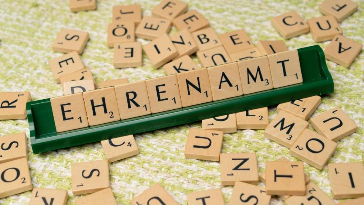 Close-up of wooden letter tiles spelling 'Ehrenamt' on a green tile holder with scattered tiles.