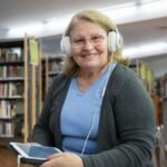 Senior woman smiling while listening to audiobooks on tablet in library setting.