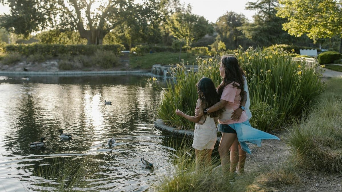 A family enjoys a tranquil afternoon in the park by a pond with ducks under soft sunlight.