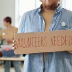 A man holds a 'Volunteers Needed' sign in a charitable organization setting, highlighting the need for help.