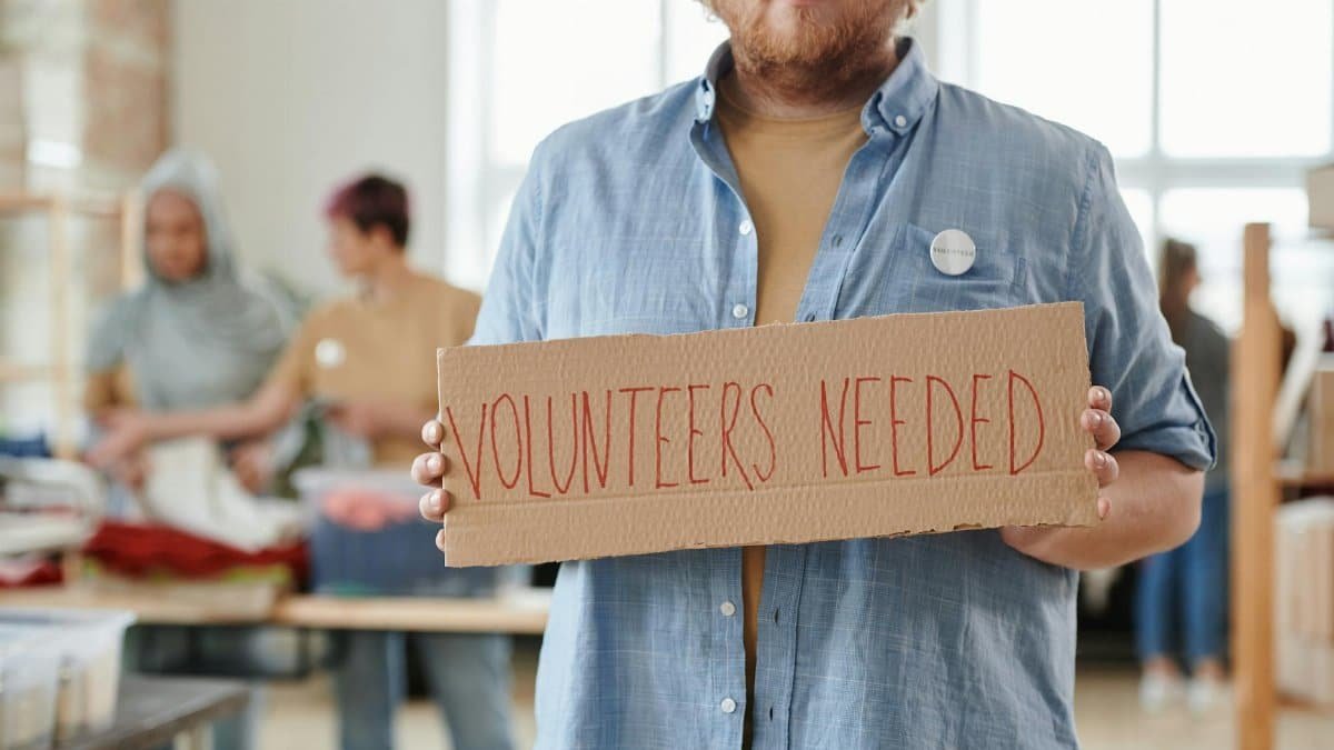 A man holds a 'Volunteers Needed' sign in a charitable organization setting, highlighting the need for help.