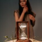 A wooden hourglass with white sand sits on a textured surface, with a woman in soft focus in the background.