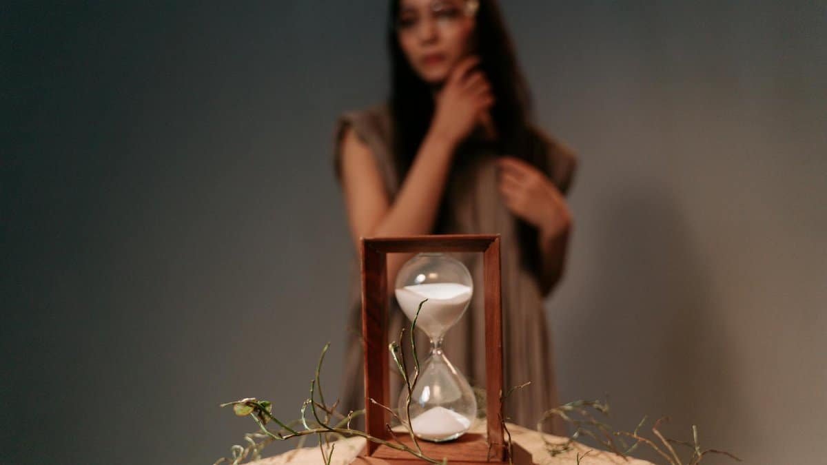 A wooden hourglass with white sand sits on a textured surface, with a woman in soft focus in the background.