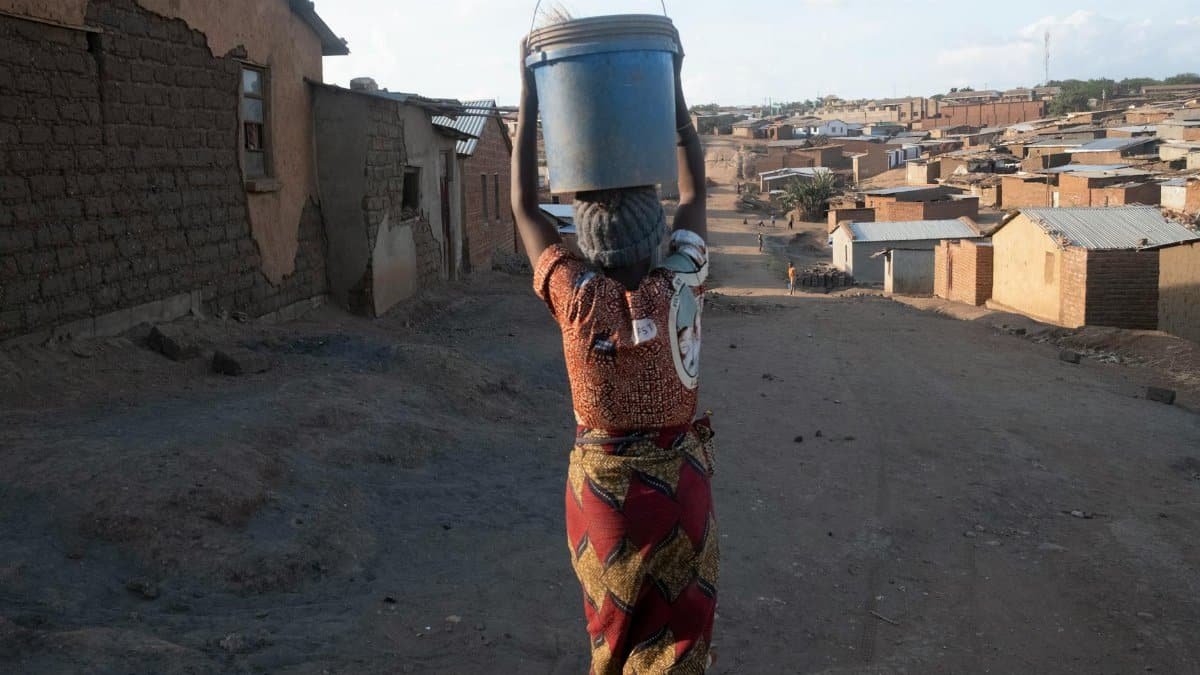 A woman balances a water bucket on her head in a rural village setting.