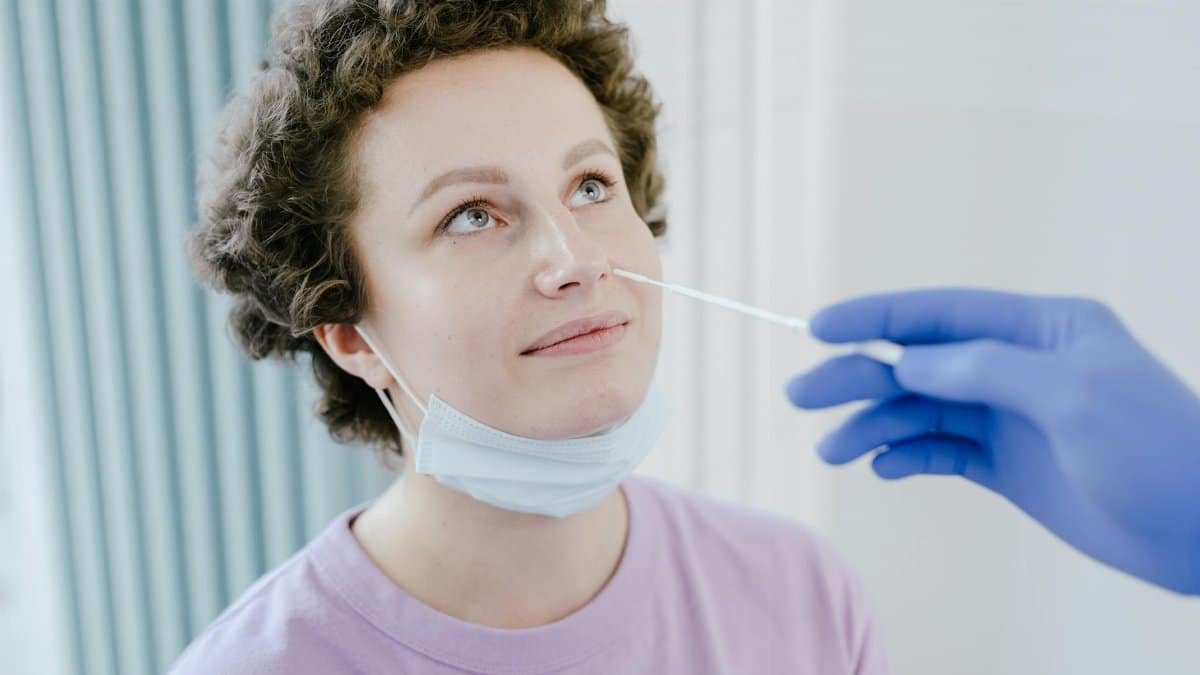 A woman in a medical facility receiving a nasal swab COVID-19 test, wearing a mask, smiling.