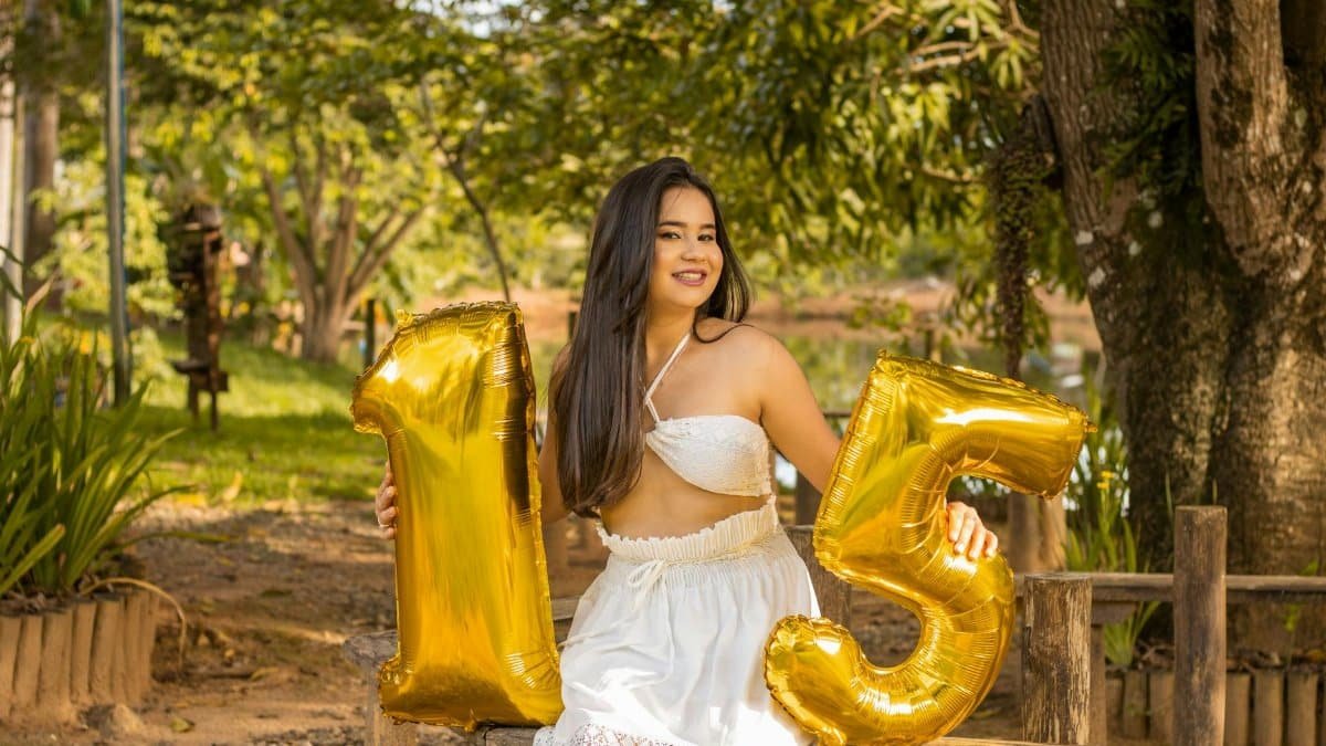 A joyful young woman in white dress celebrating her 15th birthday with balloons outdoors.