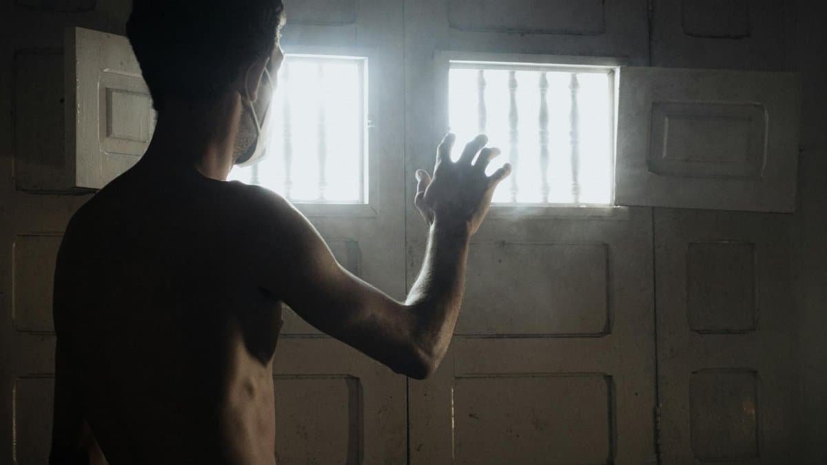 Shirtless man in a mask reaches towards light through a window, symbolizing hope.