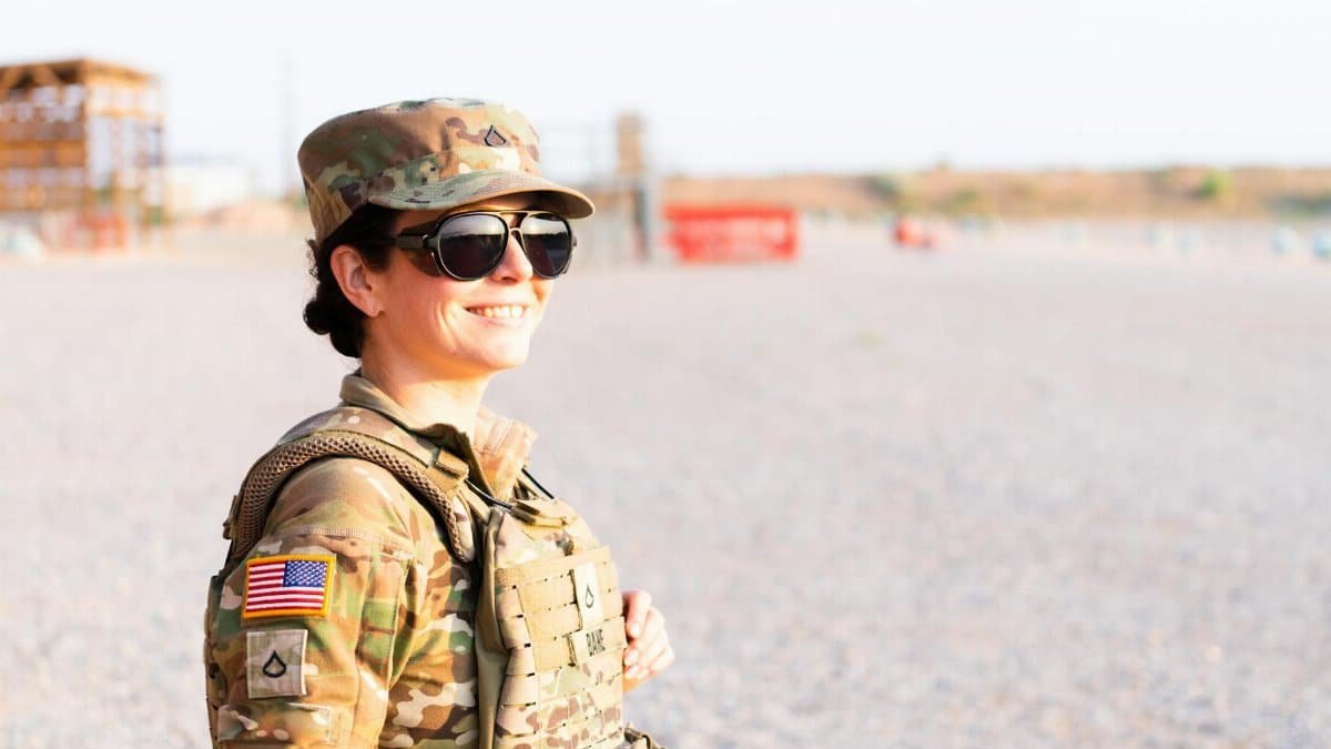 Adult female soldier in camouflage uniform smiling confidently while standing outdoors on a bright day.