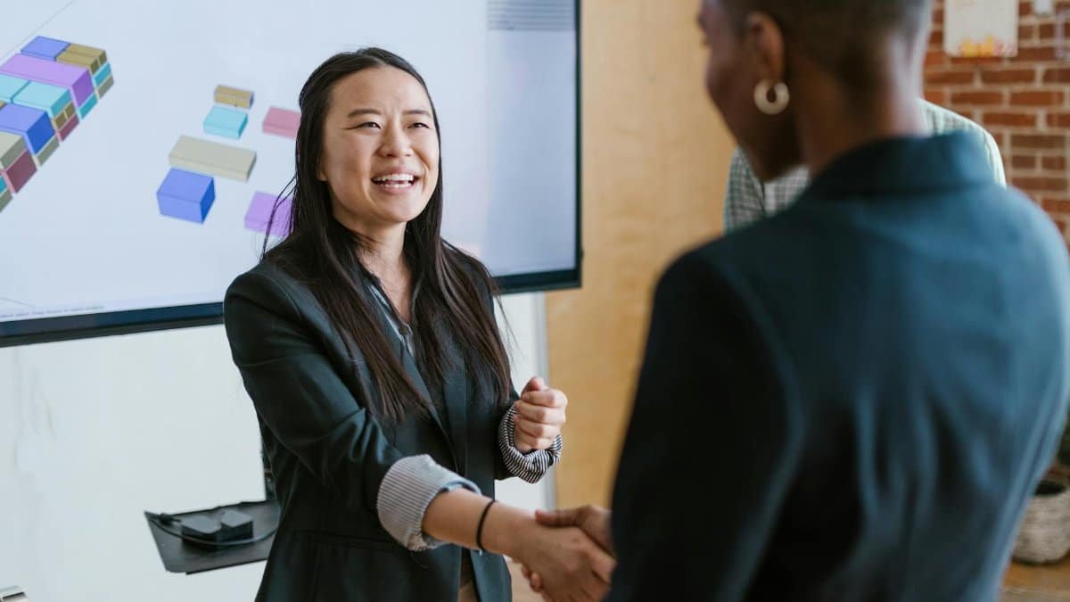 Two women shaking hands in a modern office environment with a presentation screen.