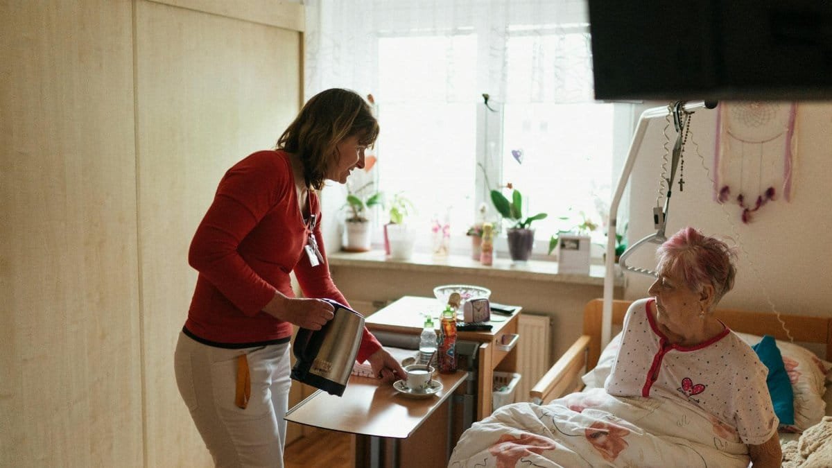 A caregiver serves tea to a senior woman in a cozy bedroom setting, fostering comfort and care.
