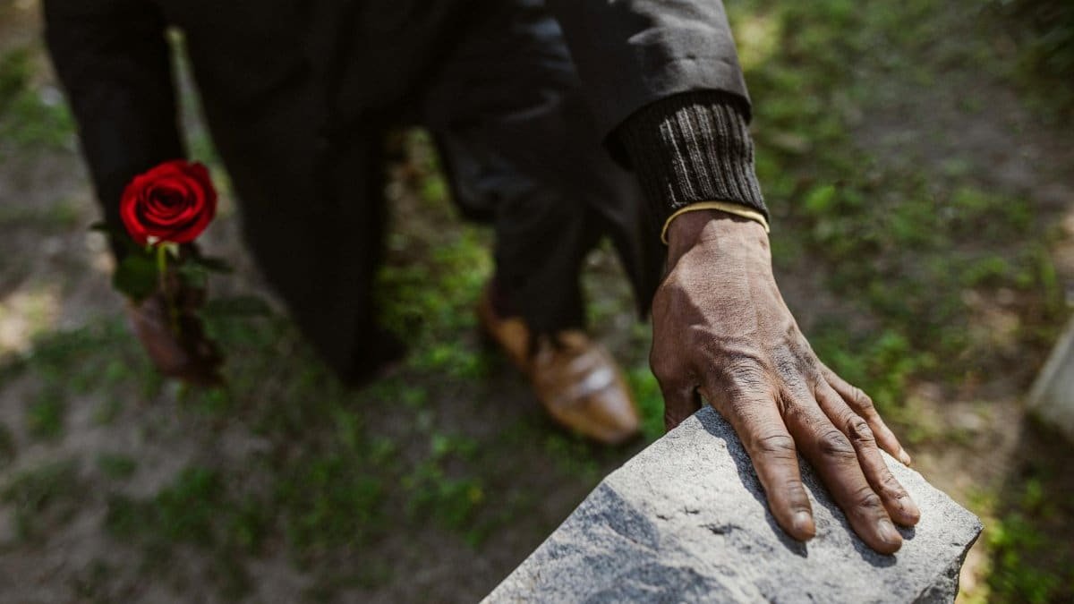 A man mourns a loved one, holding a rose by a gravestone, signifying loss and remembrance.