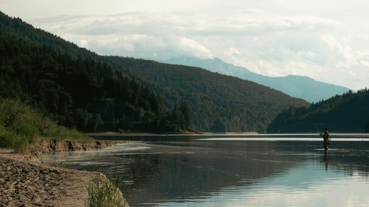 A lone fisherman fly fishing in a serene mountain lake surrounded by lush green forests during sunset.