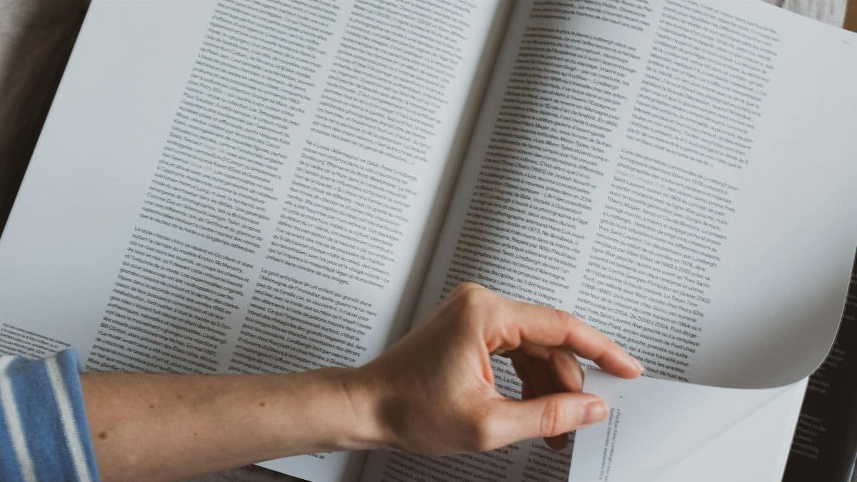 From above of crop faceless person reviewing pages of popular science book while lying on bed