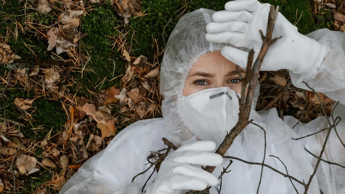 A person wearing a protective hazmat suit lies on a forest floor, holding a tree branch.
