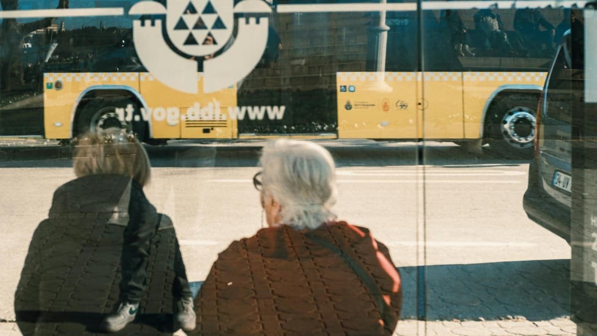 Two individuals at a bus stop with a yellow bus reflection creating an urban scene.