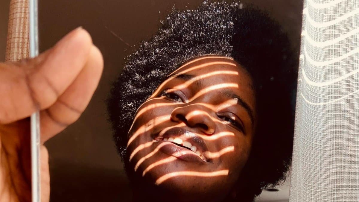 African American woman smiling in mirror reflection, sunlight creates artistic stripes on face.