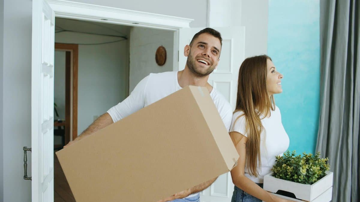 A cheerful couple moving into their new home, carrying boxes and a plant.
