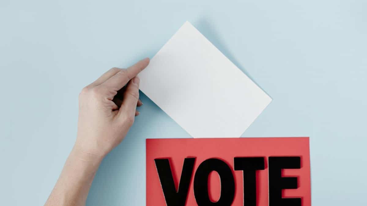 A hand places a ballot into a red box with the word VOTE against a blue background.