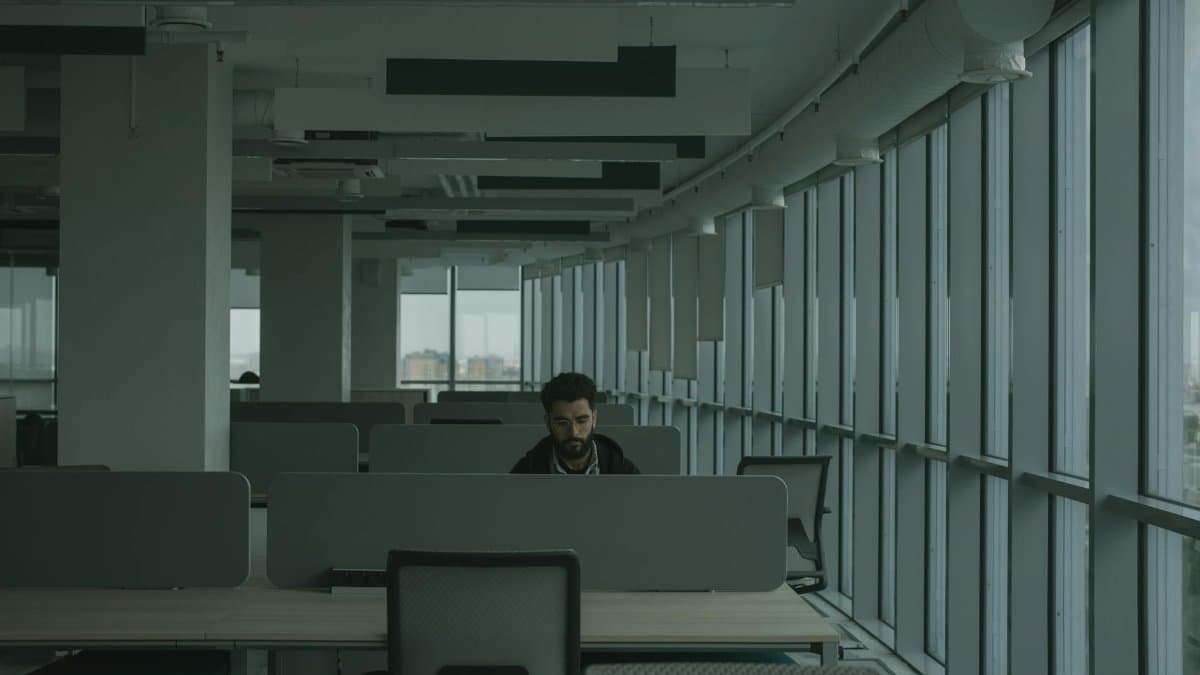 An adult man works alone in a contemporary open-plan office, surrounded by desks and dividers.