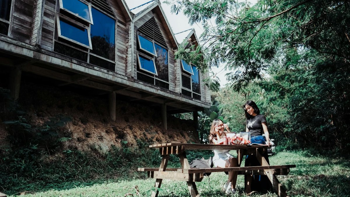Two people enjoy a picnic near a rustic wooden cabin amidst lush greenery on a sunny day.
