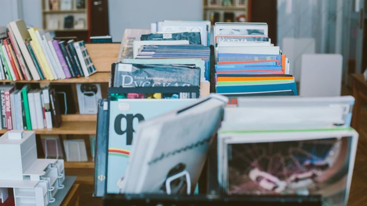 A close-up view of a bookshelf filled with various stacked books and magazines in a library setting.