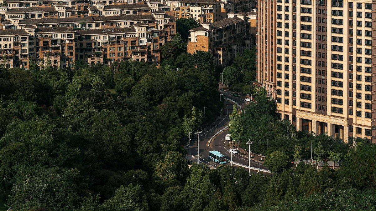 Aerial photo showcasing urban residential buildings surrounded by lush green trees, featuring a winding road below.