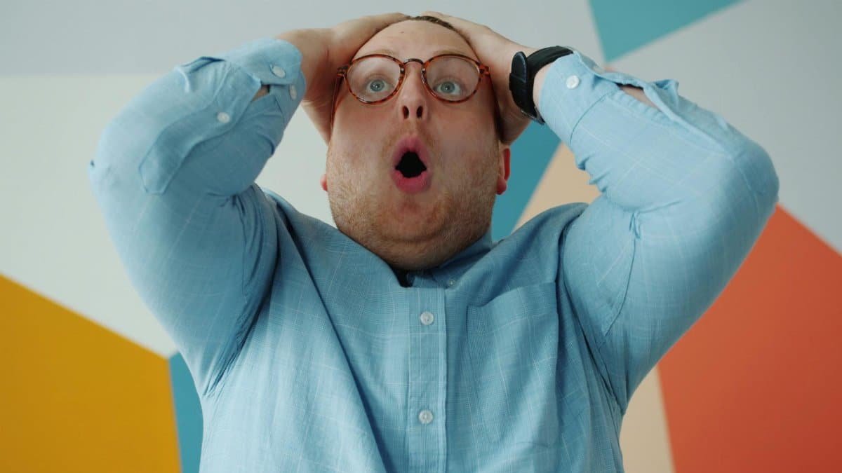 A man with round glasses expresses surprise holding his head in disbelief.