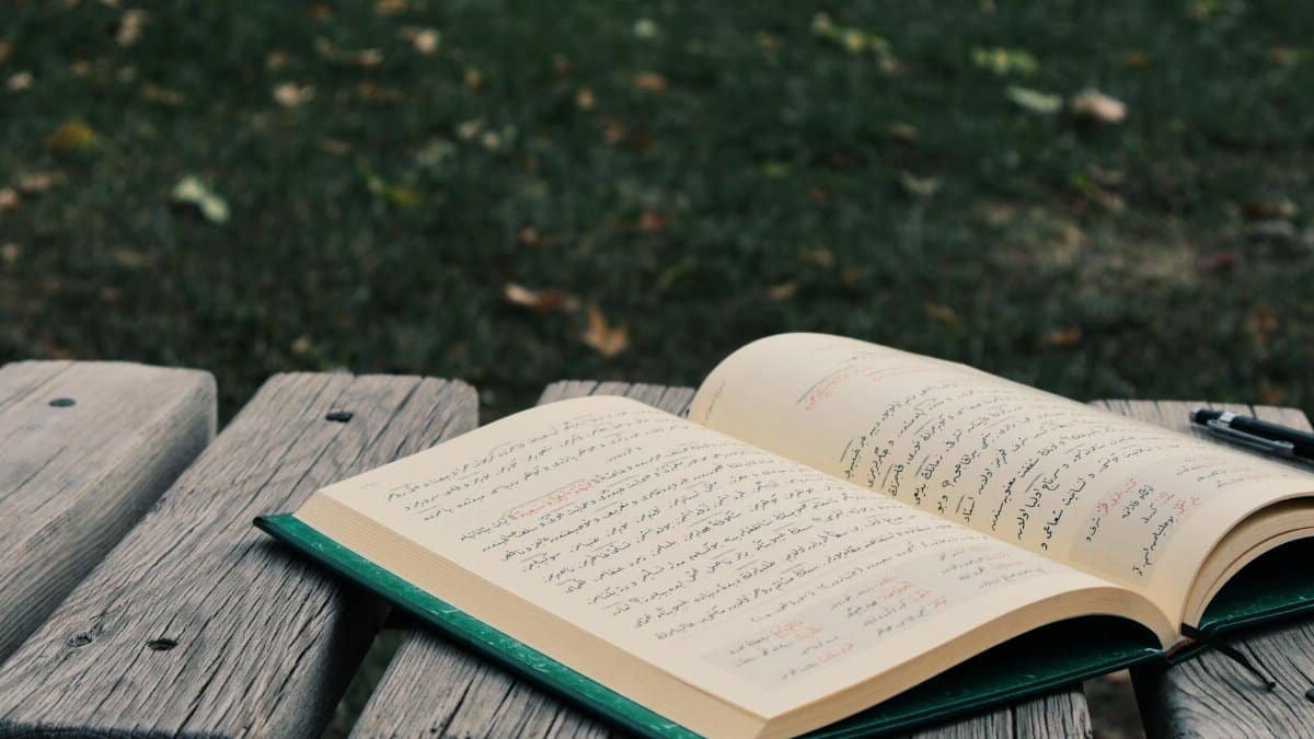 An open book with Arabic script on a wooden bench outdoors in a park.