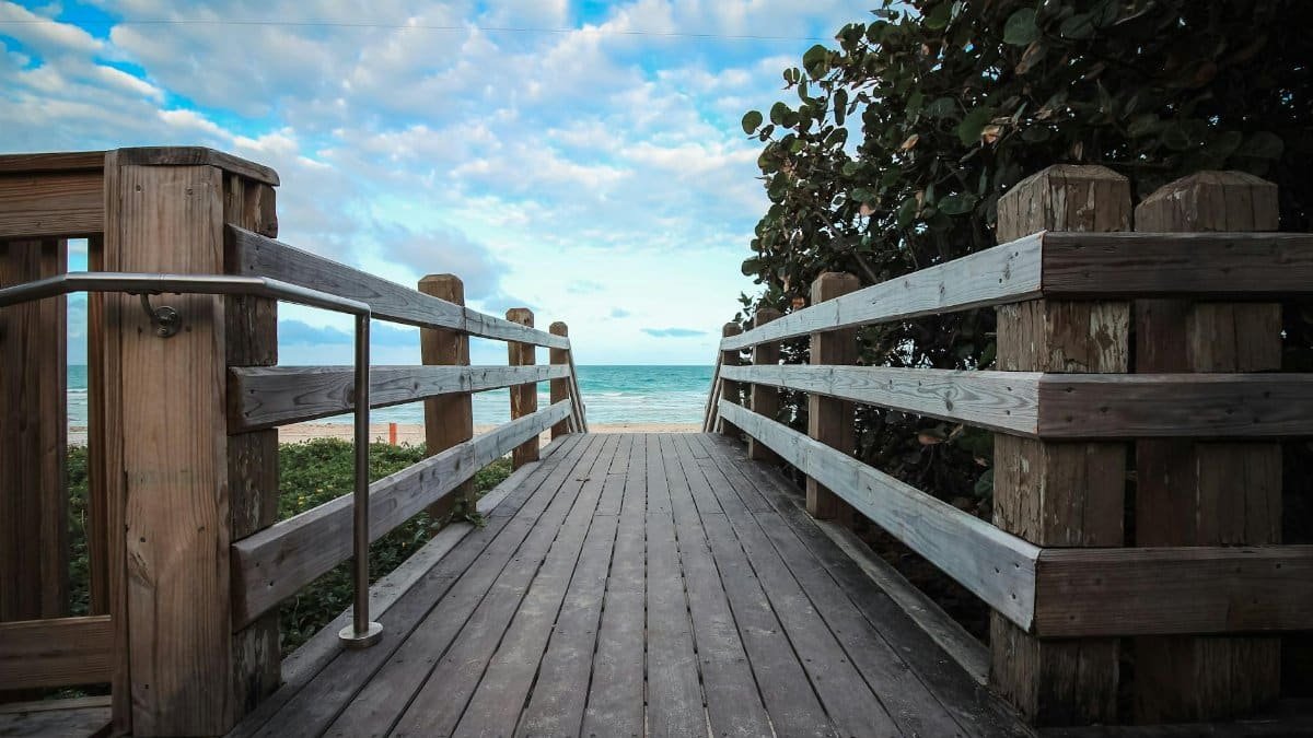 Scenic wooden boardwalk in Miami, Florida leading to a serene beach with lush greenery and blue skies.