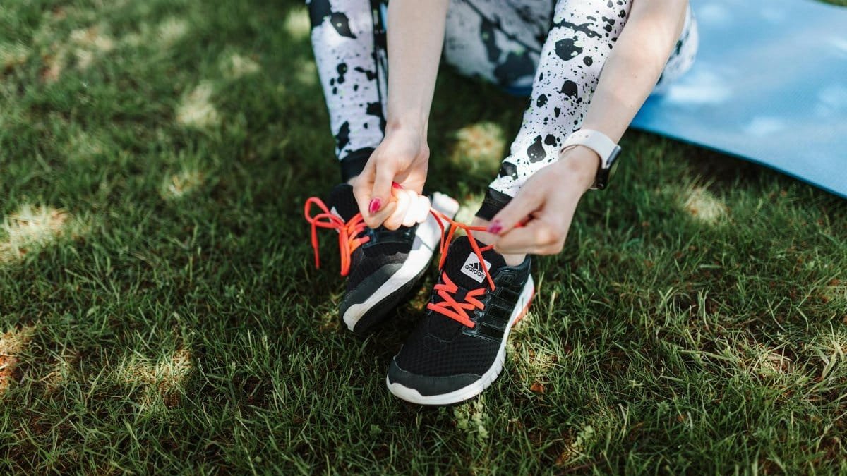 Woman tying Adidas sneakers on grass with yoga mat nearby, ready for exercise.