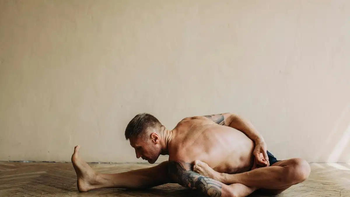 Tattooed man practicing a challenging yoga pose indoors, showcasing flexibility and strength.