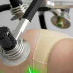 Side view of crop anonymous female in underwear lying and doing non invasive laser body treatment against light wall in clinic
