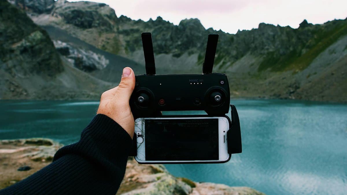 A hand holds a drone remote with a smartphone by a scenic mountain lake.