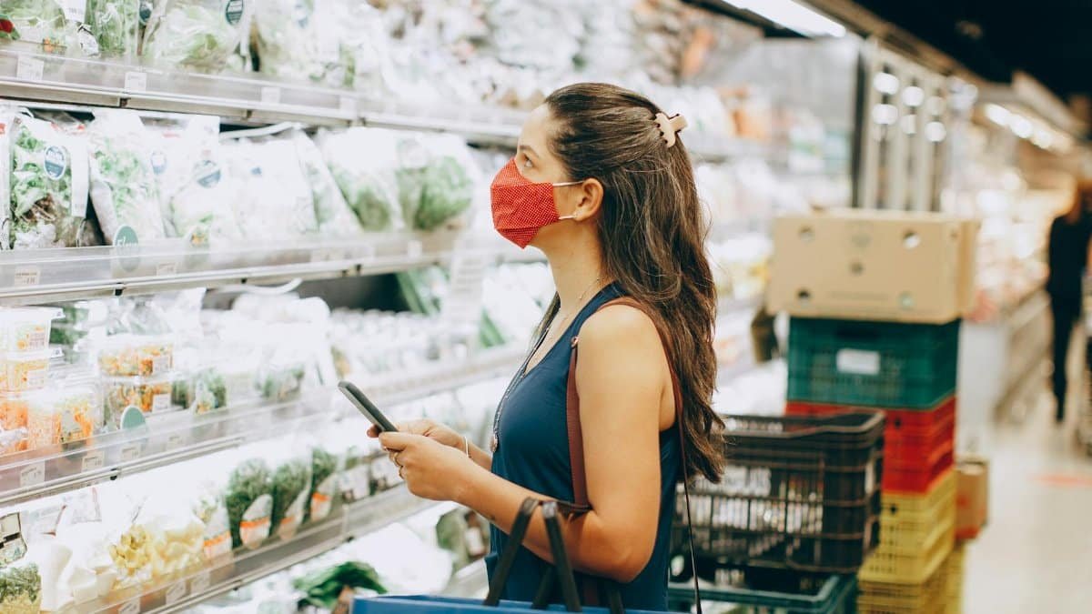 Woman with mask shopping in a grocery aisle, examining fresh produce selection.
