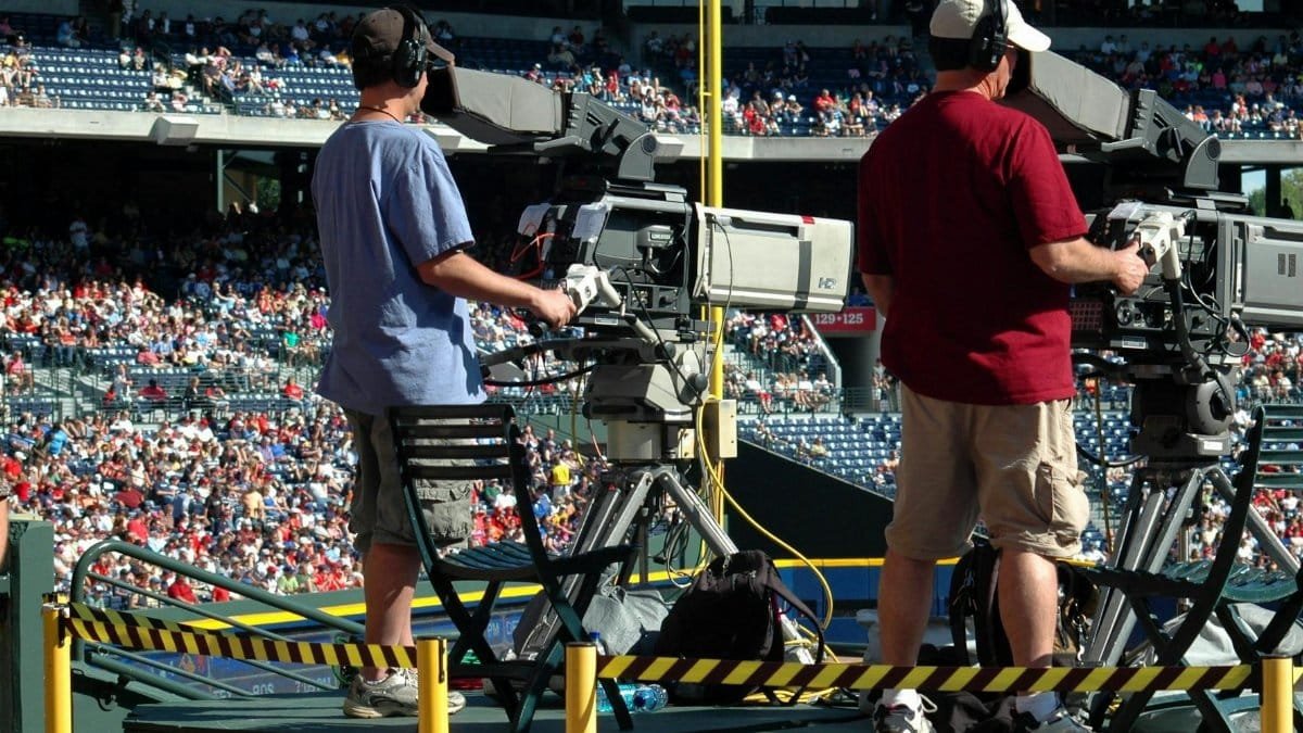 Camera operators capturing a live sports event at a crowded outdoor stadium filled with spectators.