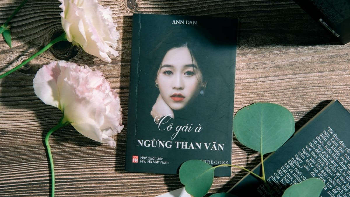A book titled 'Cô gái à' lays next to elegant flowers on a wooden table.
