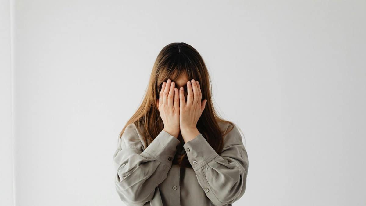 A woman with long hair stands against a white background, covering her face with her hands.