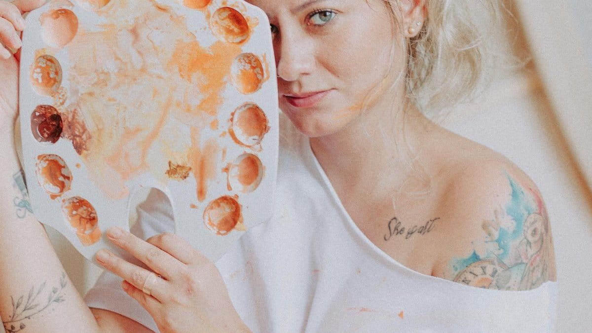 Female artist with tattoos holding a paint palette indoors, showing creativity and artistic expression.