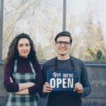 Two business owners standing outside, holding a 'Yes We're Open' sign, welcoming customers.